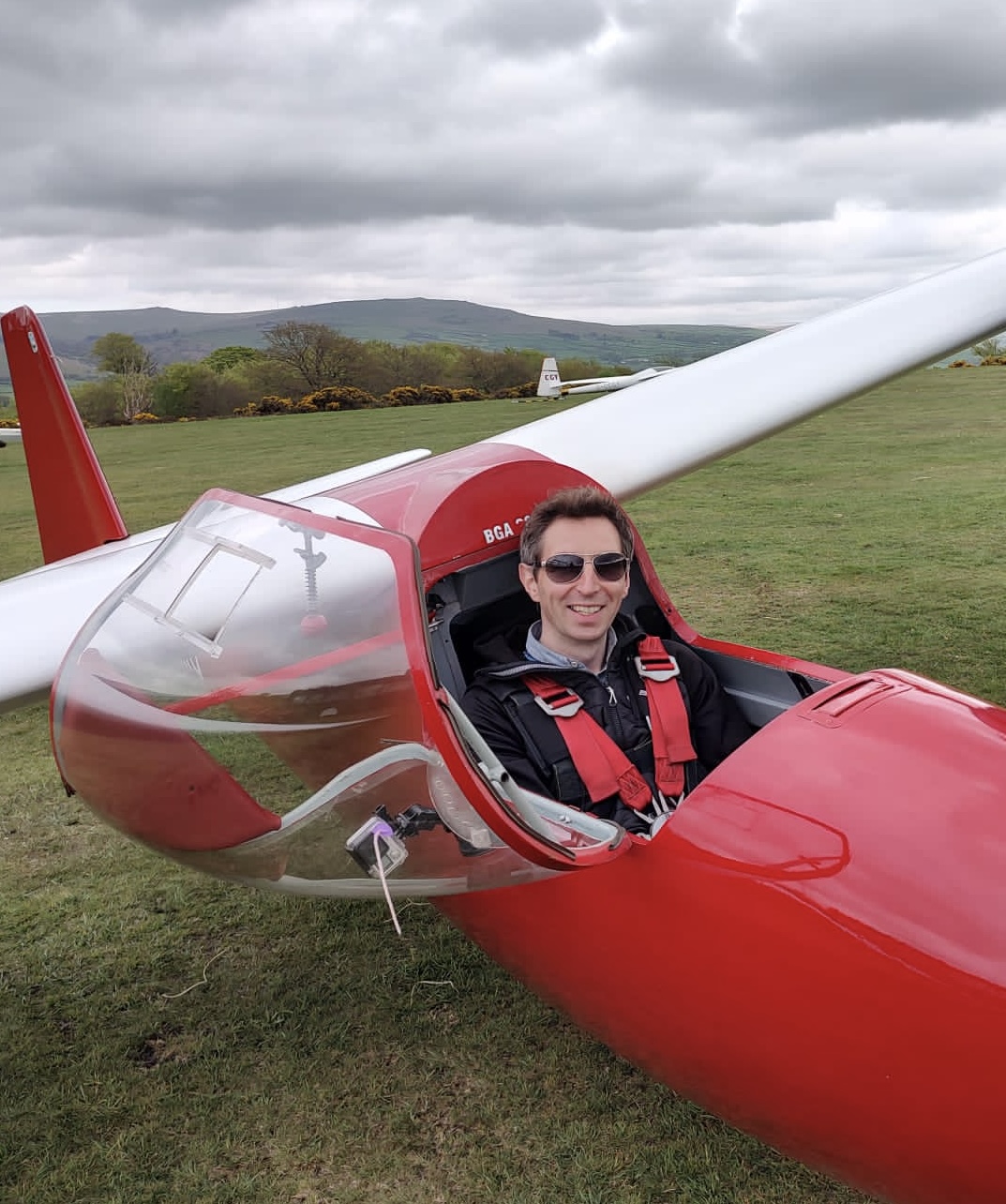 Photo of a smiling Ed Borlaise in the cockpit of a red glider with white wings on the ground