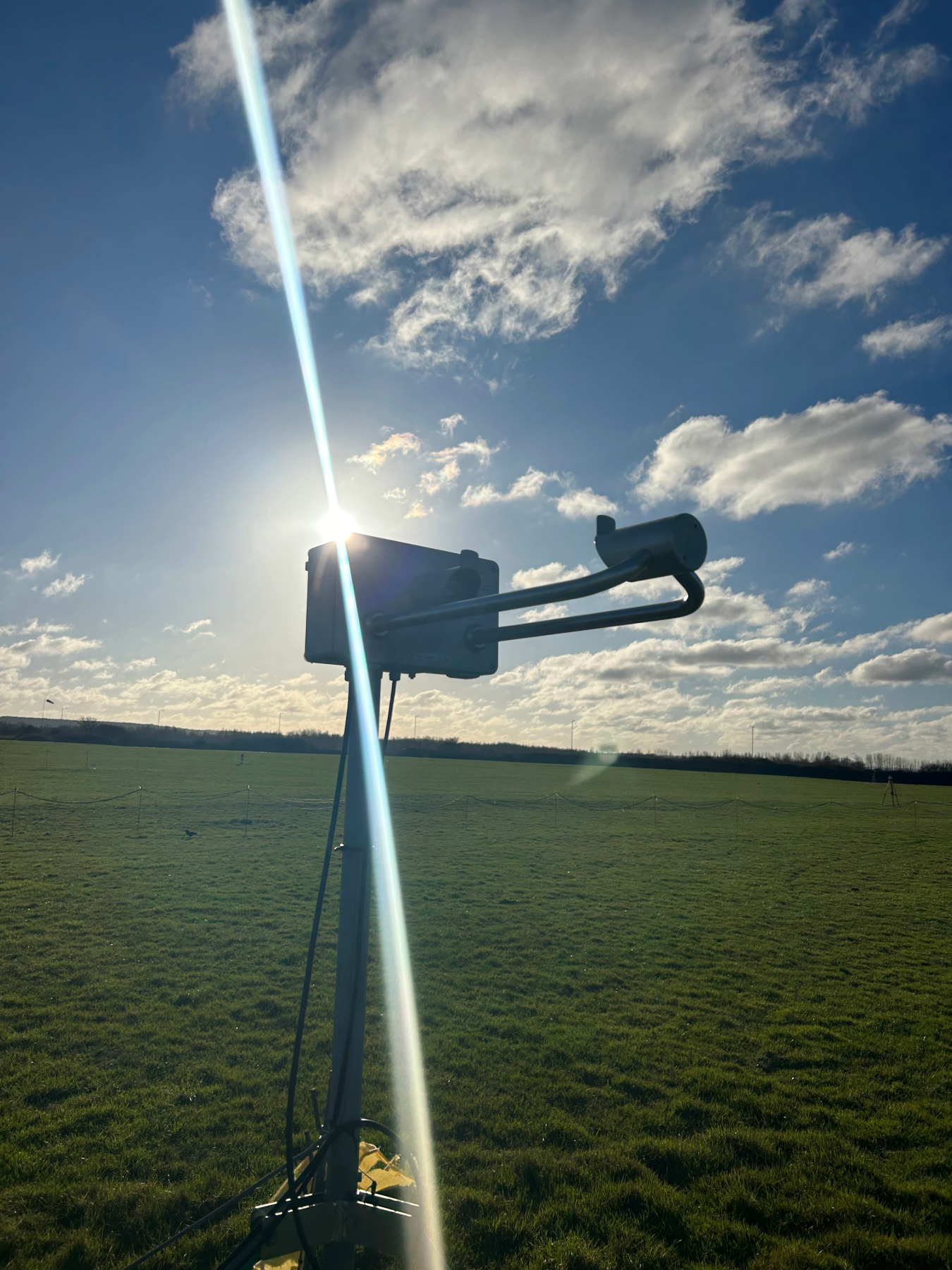 A sensor used in automated vehicles being tested in a grass field with blue skies and sunshine. The sensor is on a pole with a white box on the top.