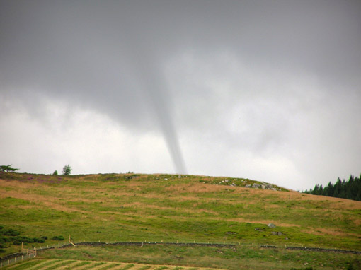 A funnel cloud forming in the UK with a grassy hill in the foreground.